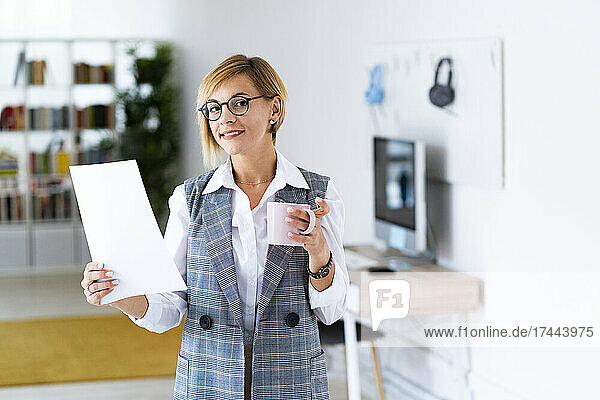 Smiling female professional holding document and coffee mug in office