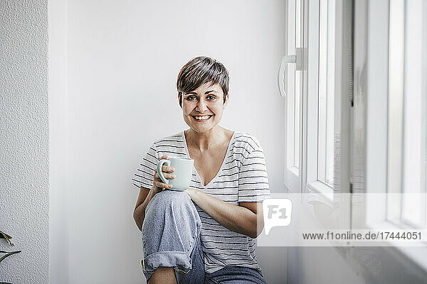Happy woman holding mug while sitting near window at home