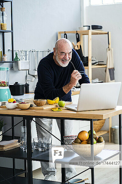 Cheerful senior man using laptop at kitchen counter