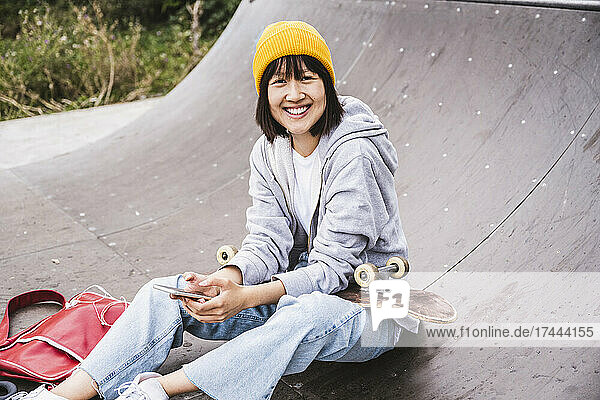 Smiling teenage girl wearing knit hat holding smart phone while sitting at skateboard park