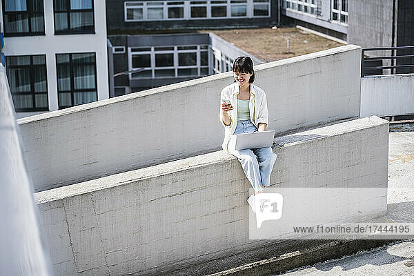 Smiling teenage girl with laptop using mobile phone while sitting on retaining wall