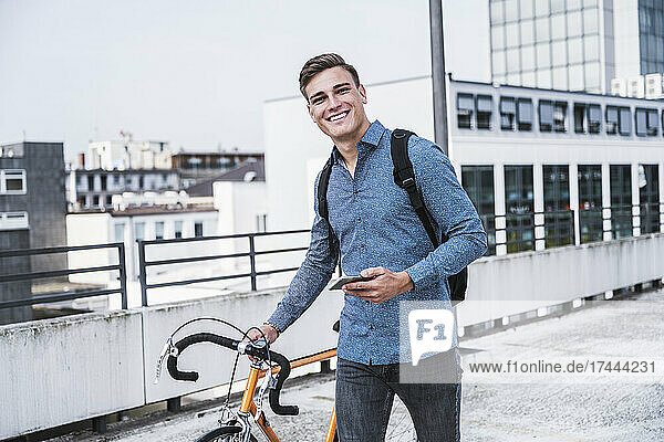 Smiling man with mobile phone and bicycle walking on rooftop