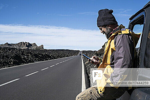 Mature man with hand in pocket using smart phone while leaning on car