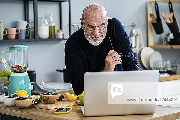 Senior man looking at laptop while leaning on kitchen counter