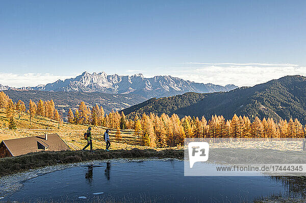 Two hikers walking past small pond in Ennstal Alps during autumn