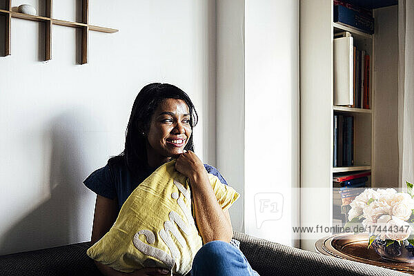 Lächelnde Frau sitzt mit Kissen auf dem Sofa im Wohnzimmer