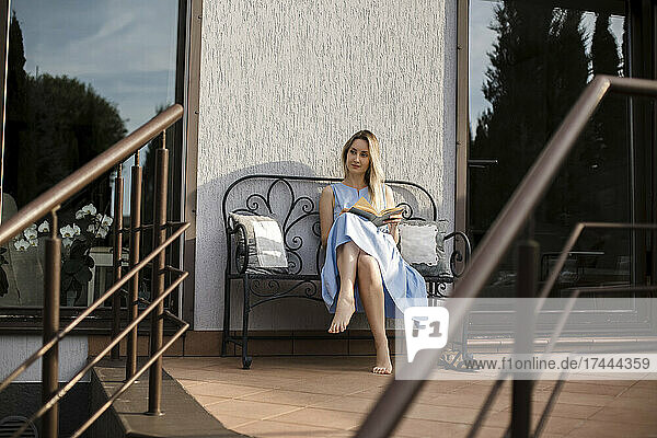 Junge Frau mit Buch sitzt auf Bank auf der Terrasse