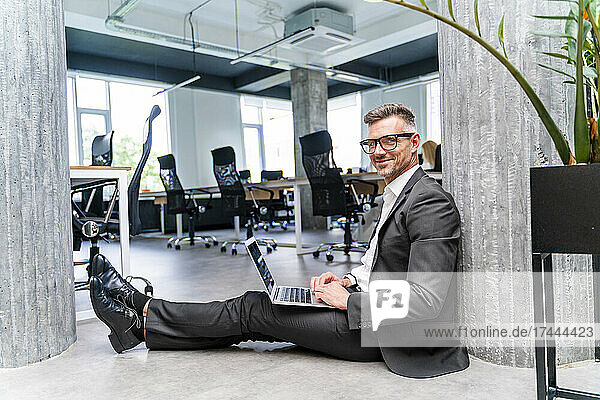 Smiling businessman with laptop sitting on floor in office