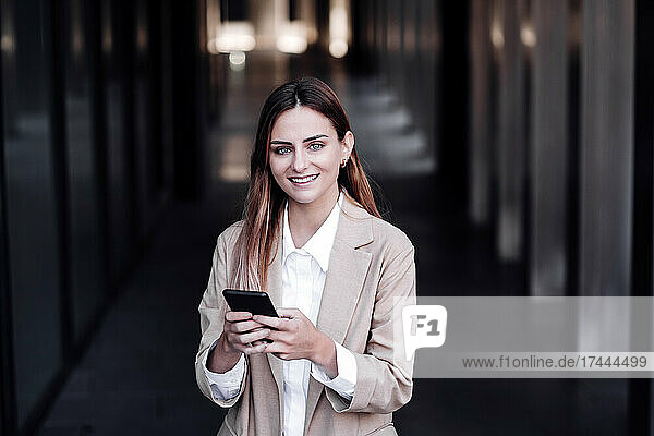 Happy female professional with smart phone standing at corridor