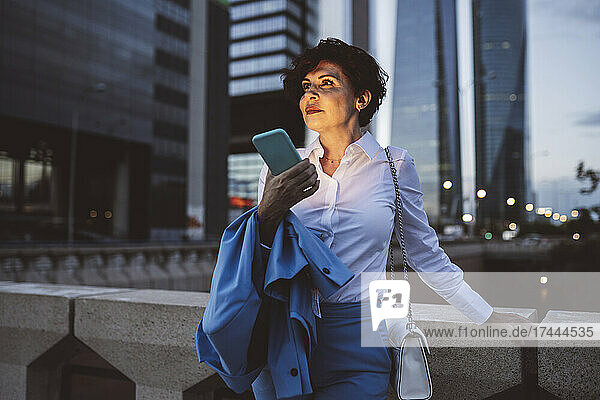 Schöne Geschäftsfrau hält ihr Mobiltelefon in der Hand  während sie in der Abenddämmerung am Geländer der Brücke steht