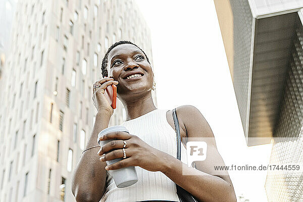 Lächelnde Geschäftsfrau mit Einwegbecher  die auf dem Smartphone spricht