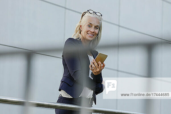 Mature businesswoman holding smart phone seen through railing
