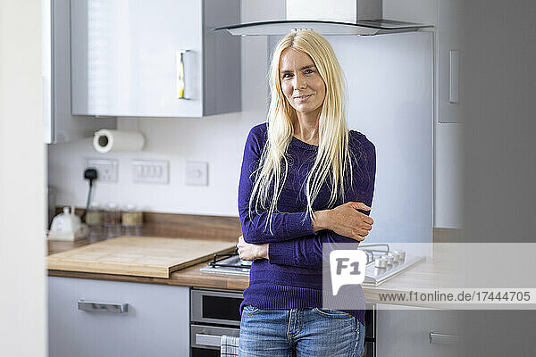 Blond woman with arms crossed leaning on kitchen counter at home