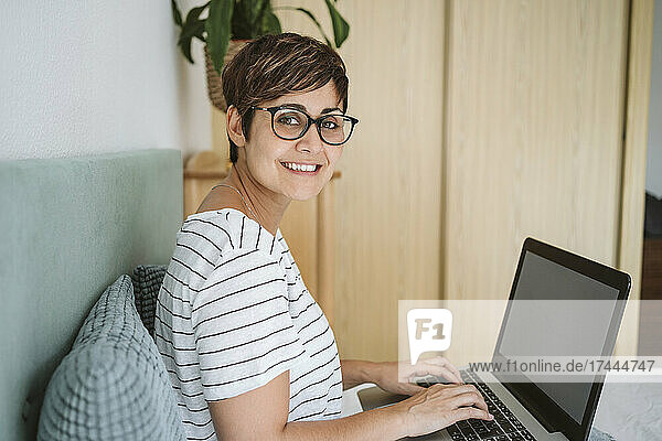Lächelnde Frau mit Brille sitzt zu Hause mit Laptop