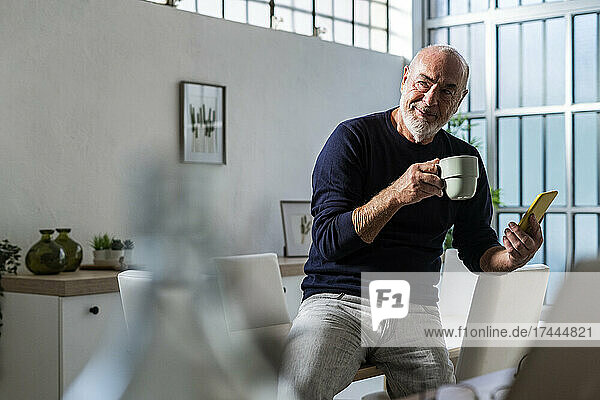 Senior man with coffee cup and smart phone sitting on table at home
