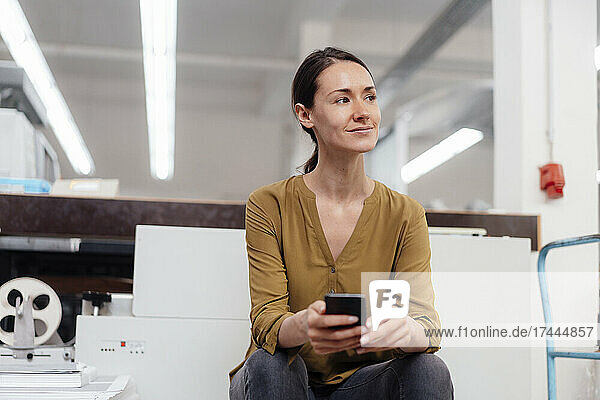 Nachdenkliche Geschäftsfrau mit Smartphone sitzt in der Werkstatt