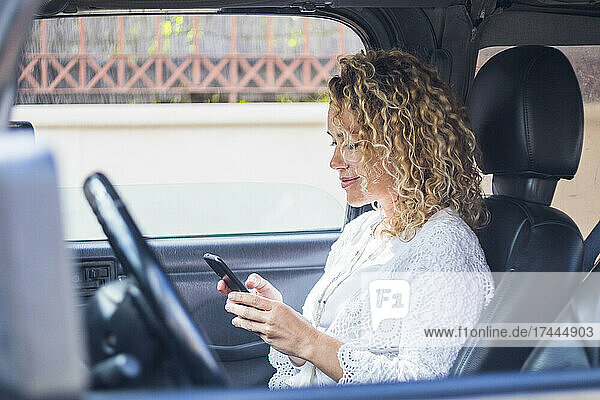 Lächelnde reife blonde Frau  die SMS über ihr Smartphone sendet  während sie im Auto sitzt