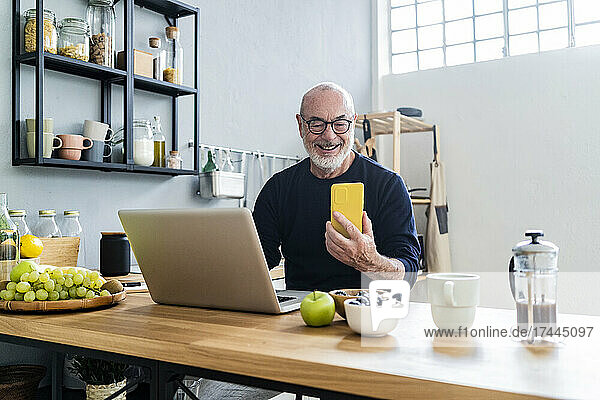 Smiling man with laptop using smart phone in kitchen