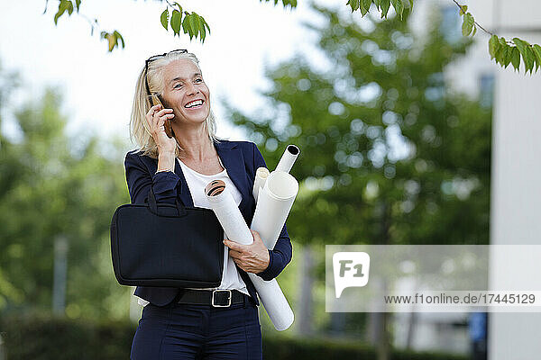 Smiling businesswoman holding blueprints while talking on smart phone