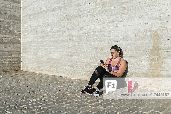 Smiling female athlete using mobile phone while sitting in front of wall during sunny day