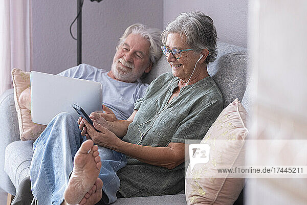 Smiling man using laptop by woman listening music on sofa in living room