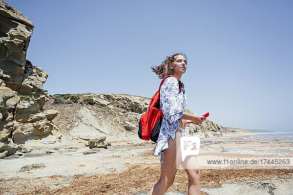 Frau trägt einen Rucksack und hält ihr Mobiltelefon in der Hand  während sie an einem sonnigen Tag am Strand spaziert