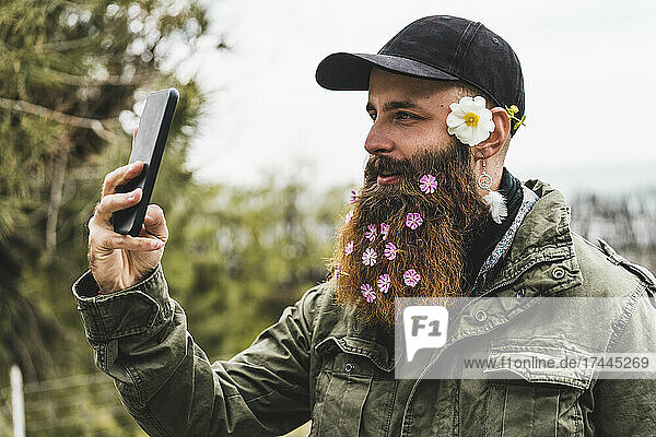 Hipster man with flowers in beard taking selfie through smart phone