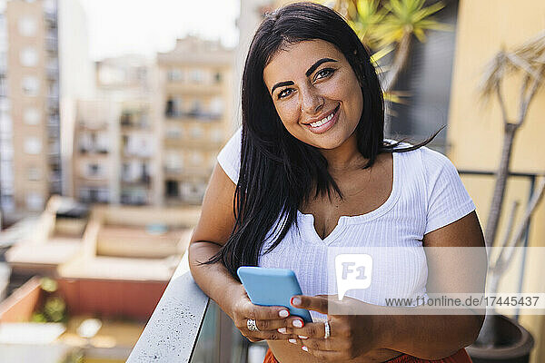Lächelnde Frau mit schwarzen Haaren  die ihr Mobiltelefon auf dem Balkon hält