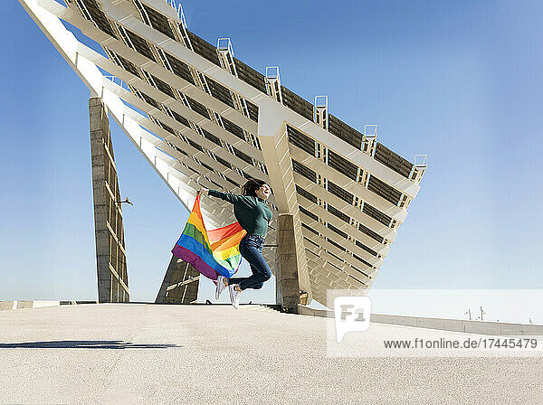 Sorglose Frau springt mit Regenbogenfahne in der Nähe von Solarenergiepanel