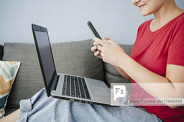Female freelancer with laptop using smart phone on sofa at home office