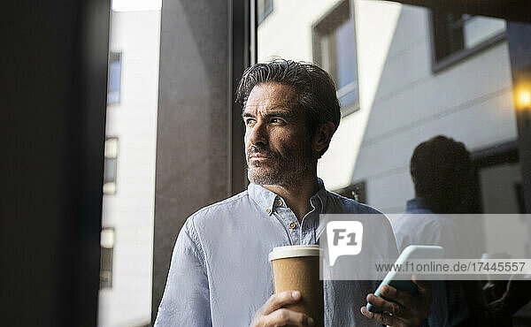 Male freelancer holding mobile phone and disposable coffee cup by window