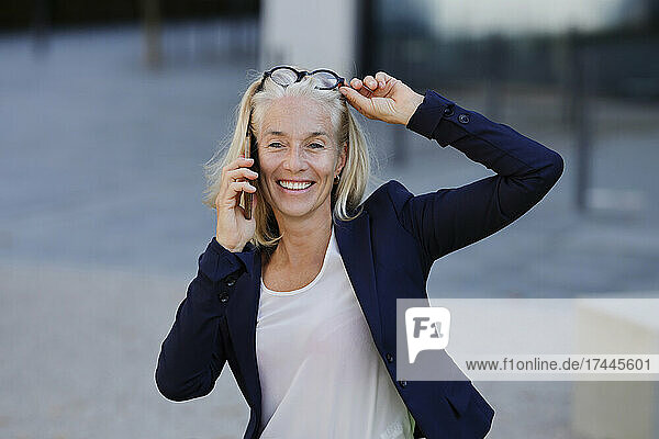 Lächelnde Geschäftsfrau  die auf dem Smartphone spricht und dabei die Brille auf dem Kopf zurechtrückt