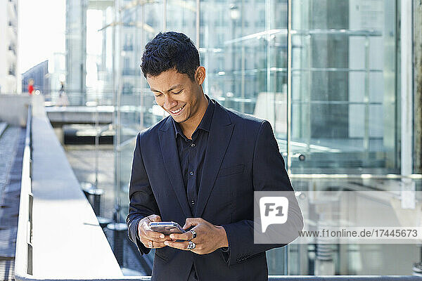 Smiling male professional using mobile phone while standing outside office building in city