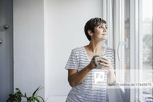 Frau hält Tasse in der Hand  während sie zu Hause durch das Fenster schaut