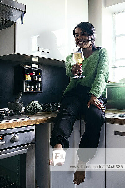 Smiling woman offering drink while sitting on kitchen counter
