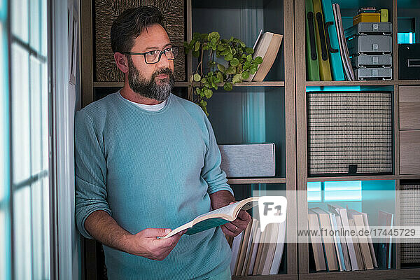 Nachdenklicher Geschäftsmann hält ein Buch in der Hand  während er sich zu Hause auf ein Bücherregal stützt