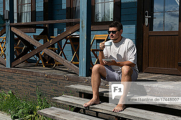 A young man is sitting on the porch of a house with a cup of coffee