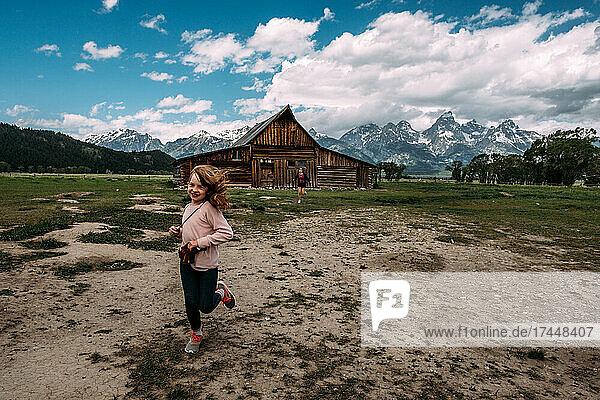 Young girls running outside an old barn near epic mountain range