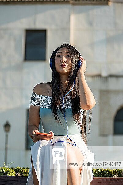 Asian girl sitting on a bench. Listening to music with headphones.