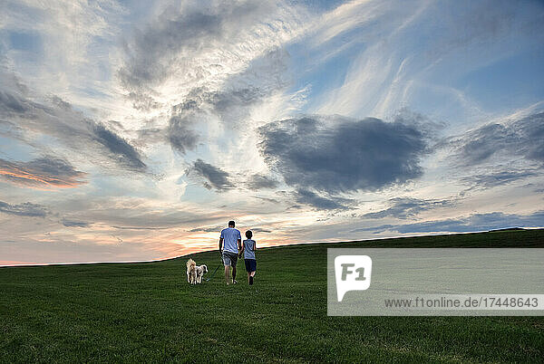 Father and son walking a dog up a grassy hill together at sunset.