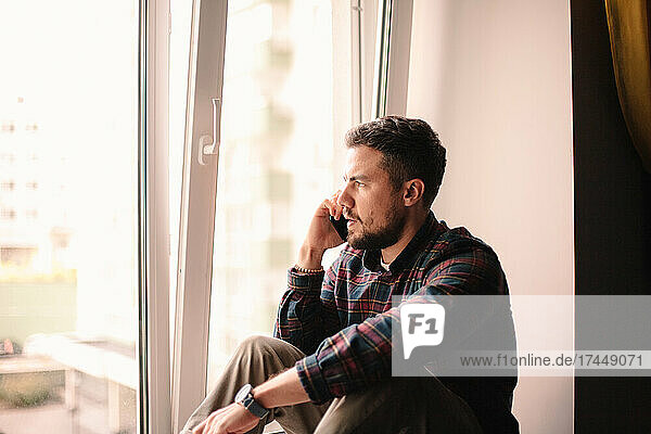 Serious man talking on smart phone sitting by window at home