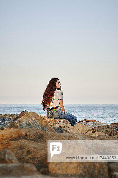 Young redhead woman looks out to sea while she is sitting on rocks