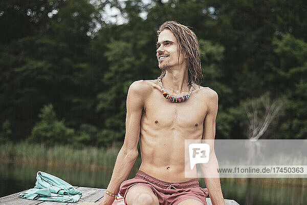 Pretty queer man smiles topless looking out at beautiful natural lake