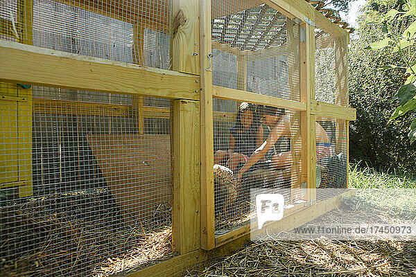 two children sit together inside enclosed run playing with chickens