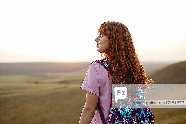 Young redhead woman with backpack looks away in sunset