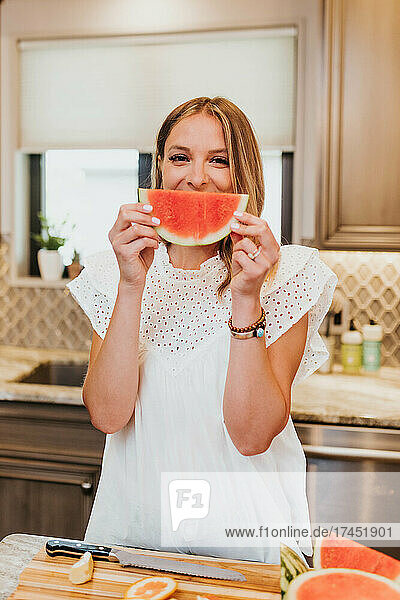 Woman holding watermelon up to her face and smiling in her kitchen