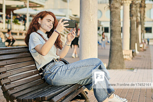Young redhead woman takes a picture with her mobile sitting in a bench