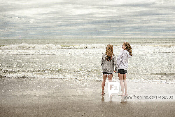 Teen girls in hoodies talk and laugh on windy Florida beach in winter.