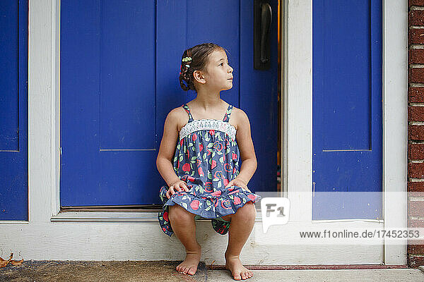 Cute little girl with braids sits barefoot in sundress on front stoop