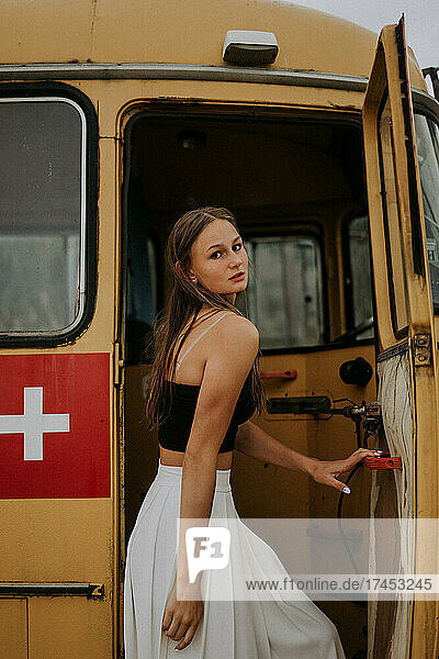 A teenager girl in a white skirt is standing by a yellow bus.